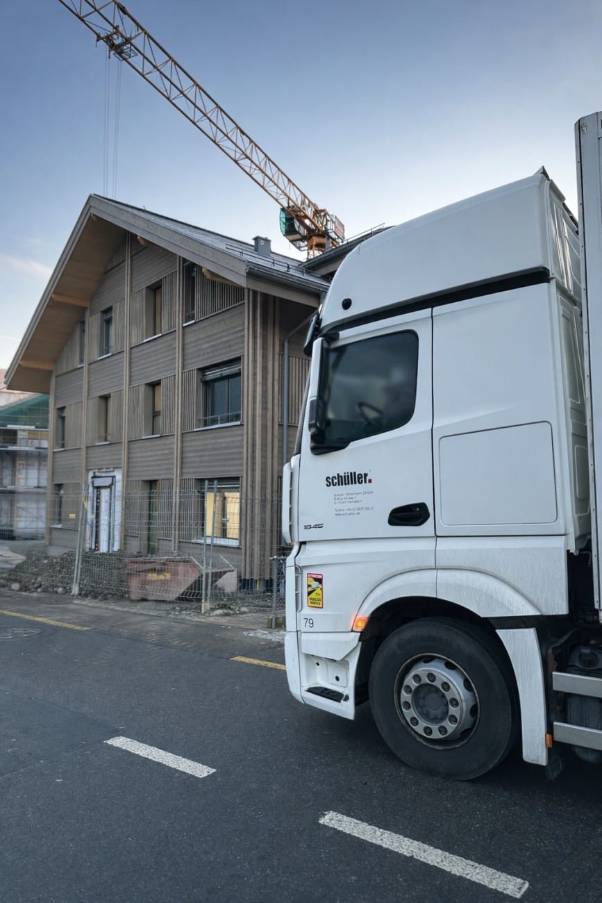 LKW vor einem Holzhaus im Bau mit Kran im Hintergrund, symbolisiert modernes Bauwesen und Logistik.