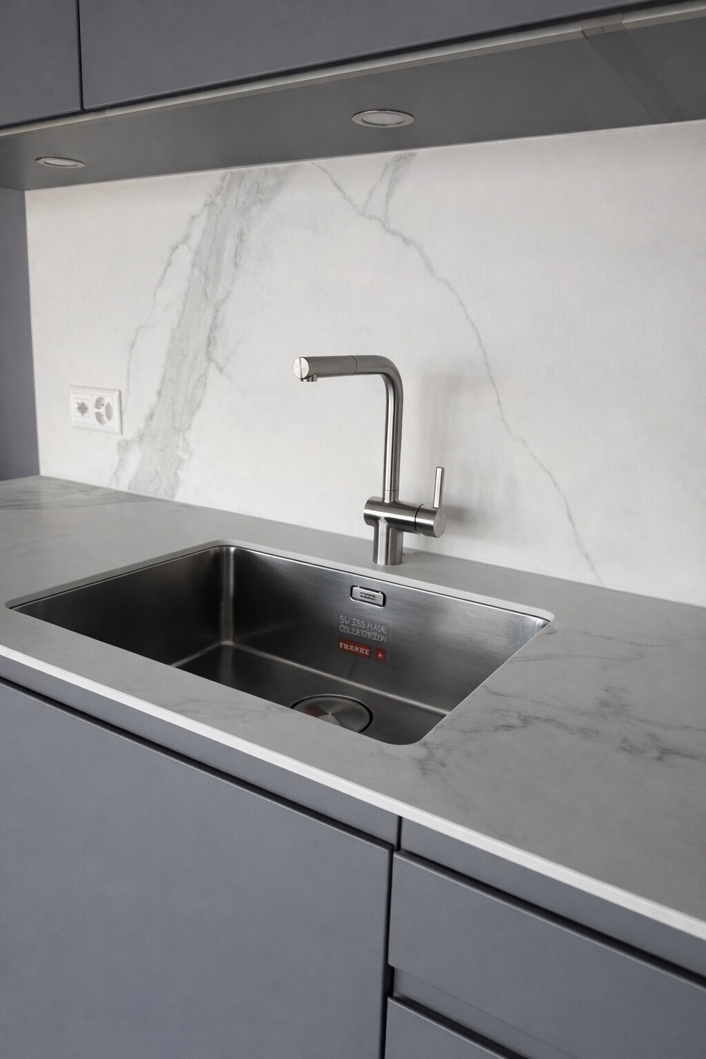 Moderne Küche mit Edelstahl-Spüle und Wasserhahn, graue Oberflächen und minimalistische Marmor-Rückwand.