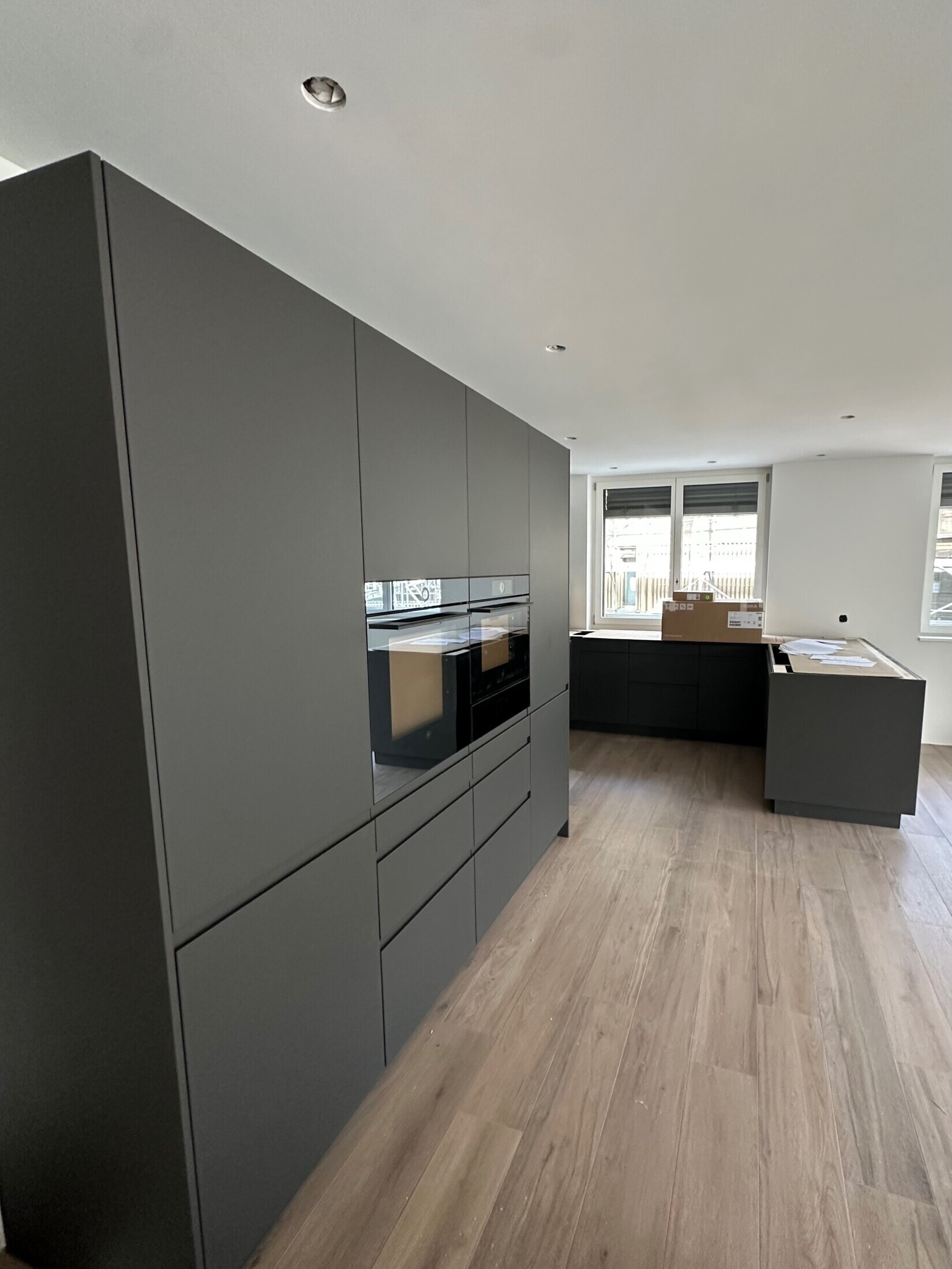 Moderne graue Küche mit Einbaugeräten und Holzfußboden, hell beleuchtet durch große Fenster.