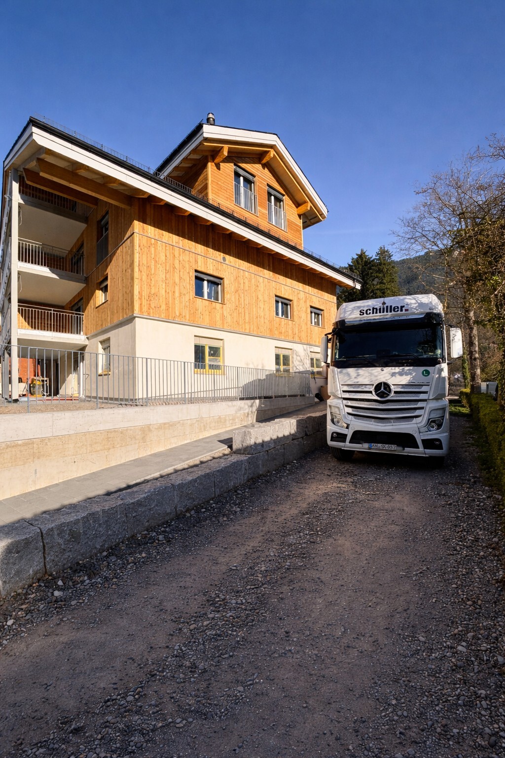 Moderner Holzbau mit LKW von Schiiler auf Schotterstraße, bei sonnigem Wetter und blauen Himmel im Hintergrund.