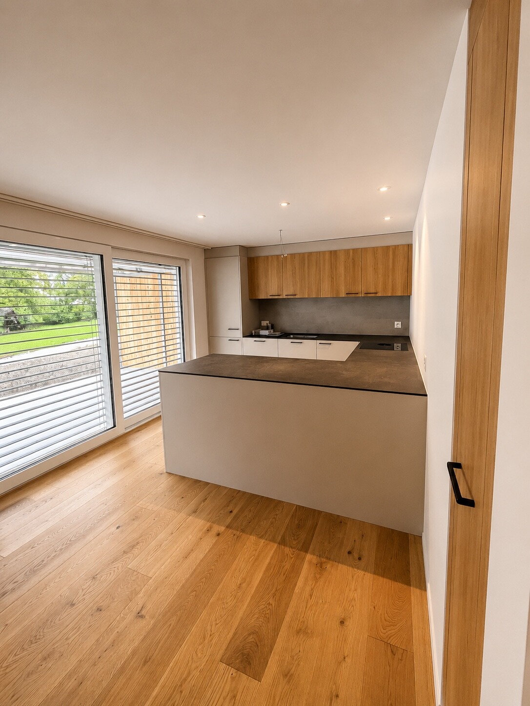 Moderne Küche mit Holzfußboden, weißen Schränken, Arbeitsplatte aus Beton und großen Fenstern mit Blick ins Grüne.